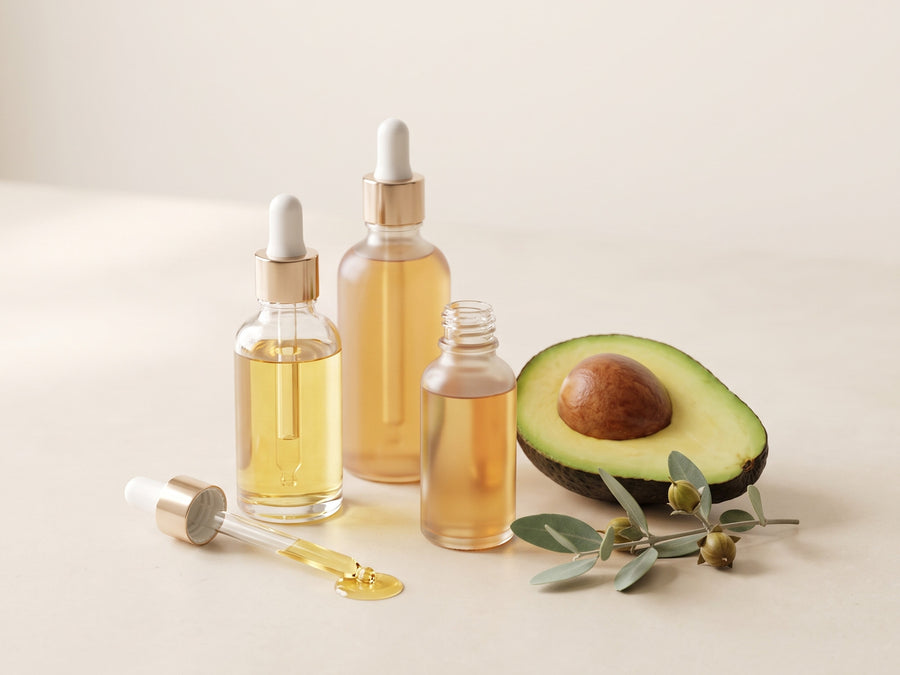 Small glass droppers of golden plant oils beside jojoba leaves and an avocado half
